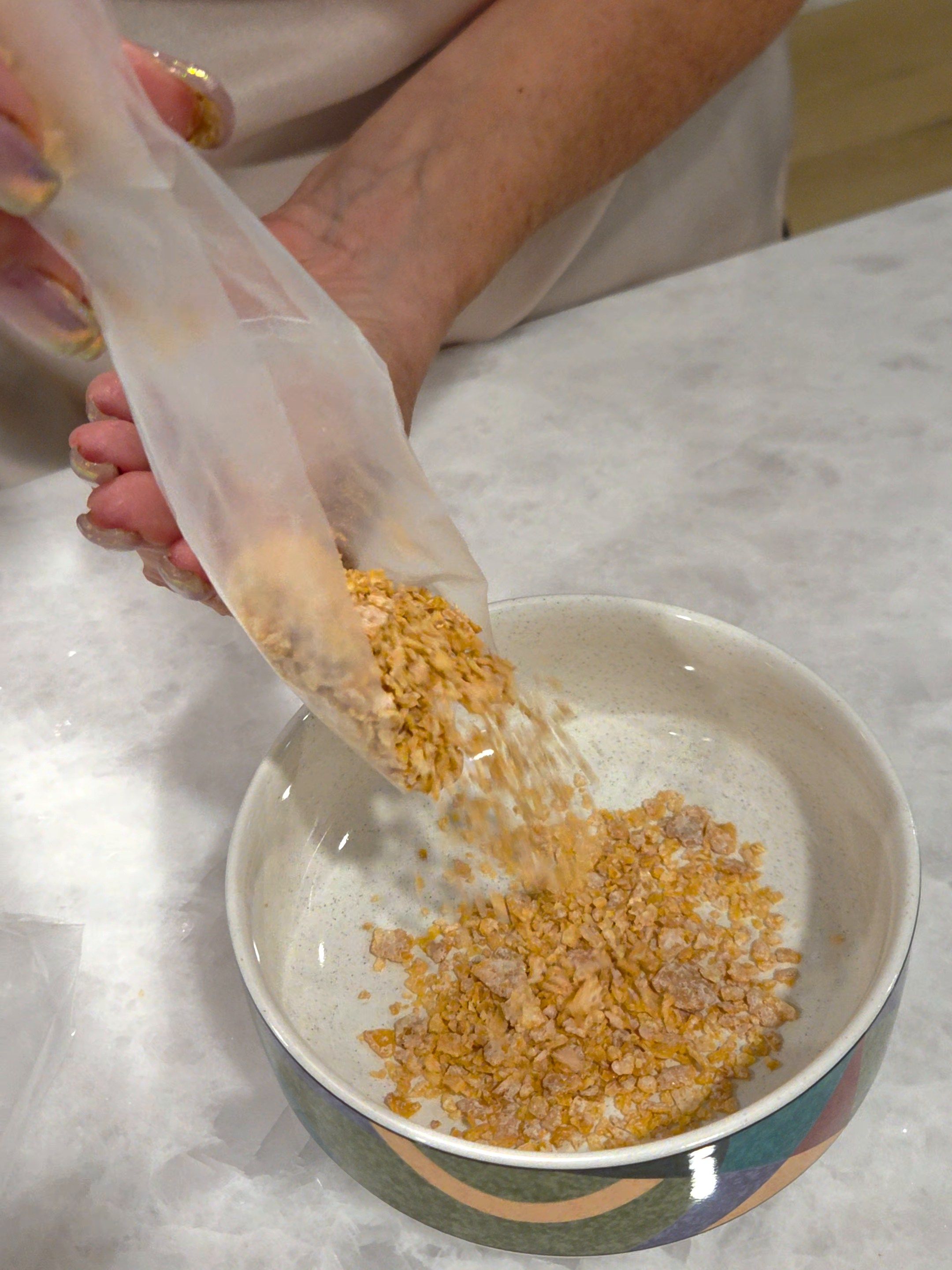 Hands pour crumbs from the small clear crumb compartment into a ceramic bowl on a marble counter.